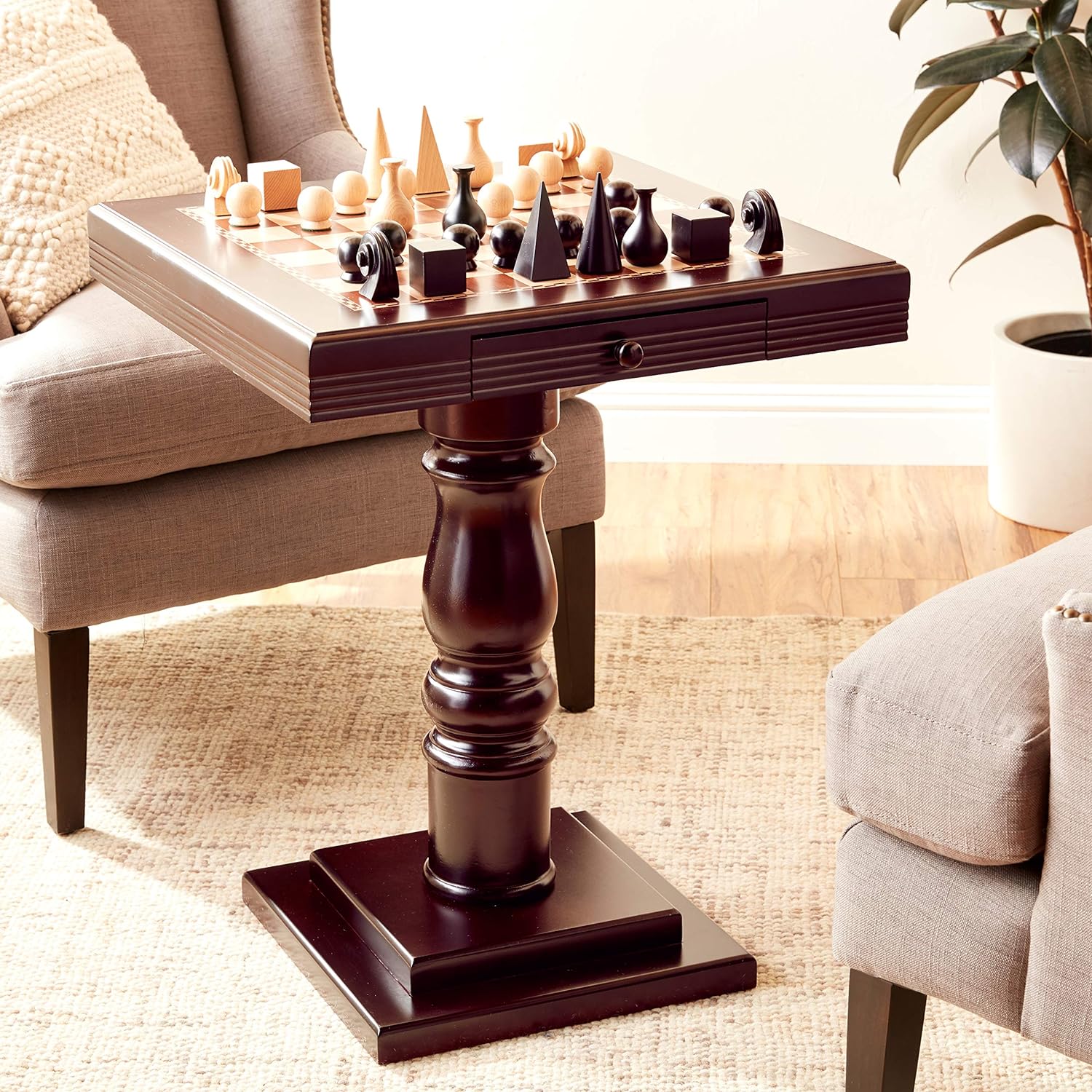 A mahogany chess table with a tournament-sized chessboard on top, featuring a pedestal base and two doors. The table is placed in a room with a chair and a plant in the background.